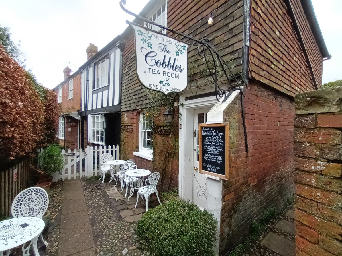 A cobbled lane with The Cobbles Tea Room hanging sign and outdoor tables